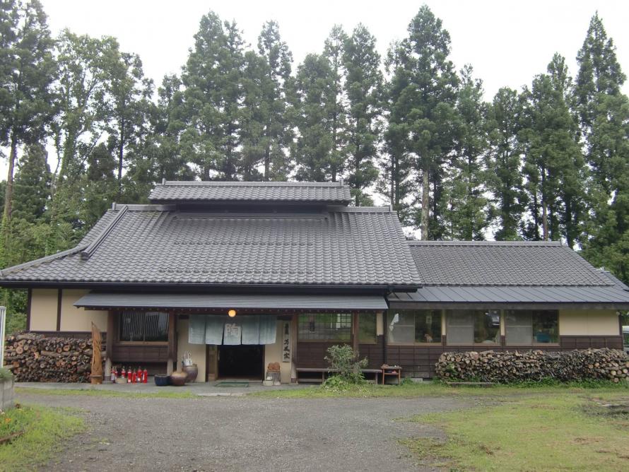蔵王町の陶芸工房「蔵王焼万風窯」の趣ある日本家屋の外観と、壁沿いに積み上げられたたくさんの薪